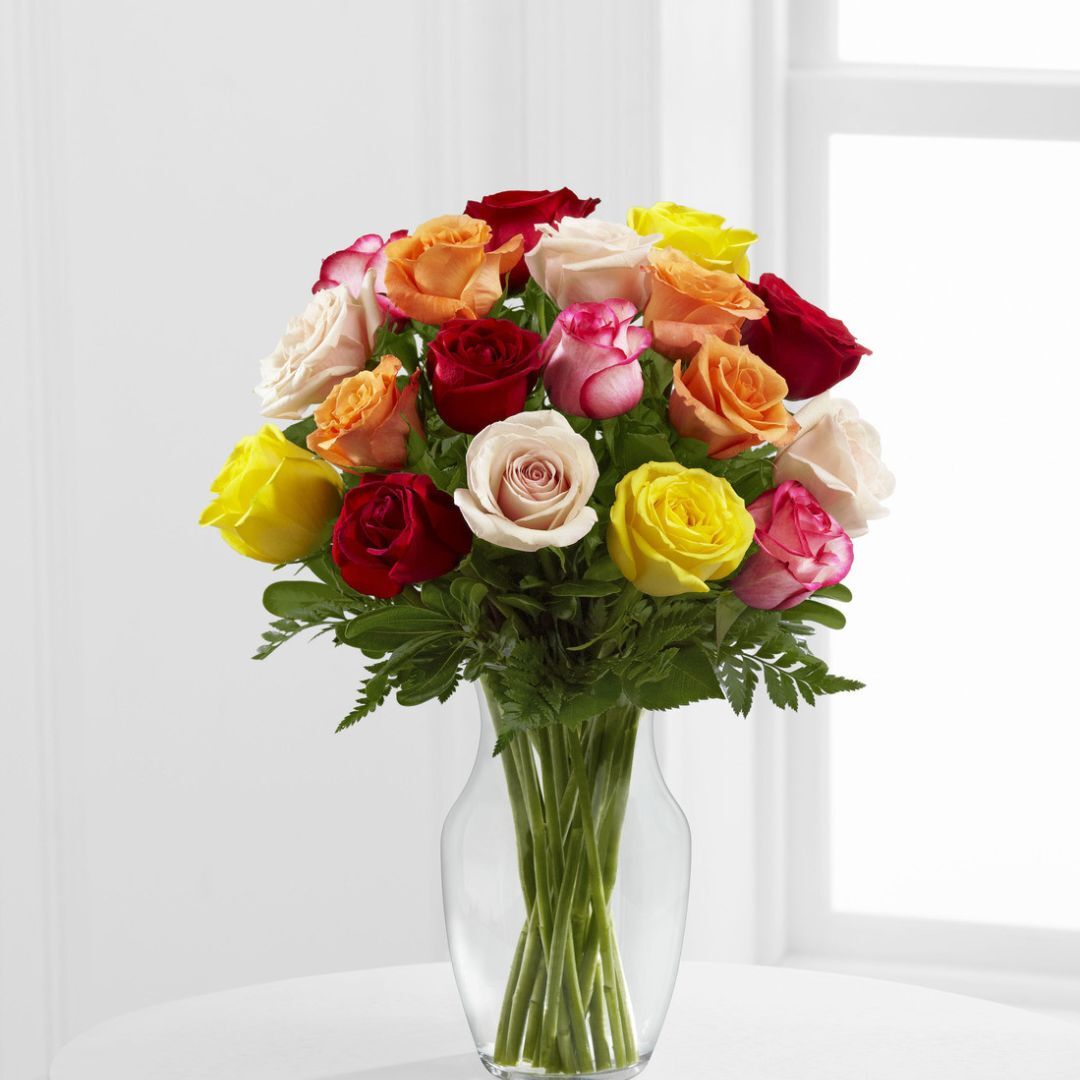 Vibrant rose bouquet with red, pink, yellow, and peach blooms in a clear glass vase, placed near a bright window.