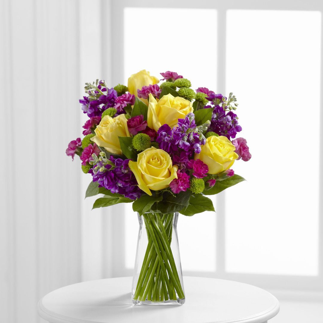 A bouquet of yellow roses, purple flowers, and greenery arranged in a clear glass vase on a white round table.
