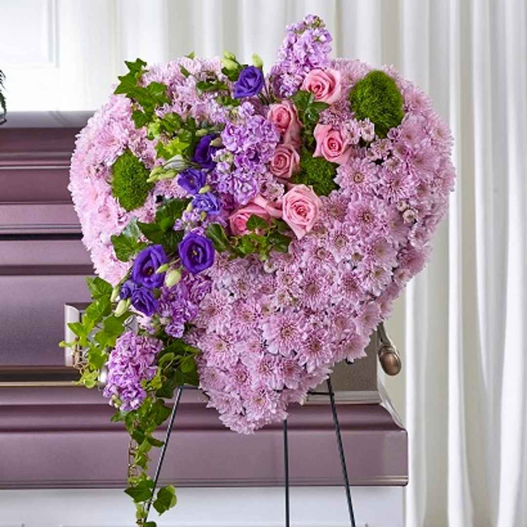 Heart-shaped funeral flower arrangement with pink, purple, and green flowers on a stand by a casket.