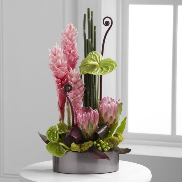 Tropical flower display featuring pink and green blooms, curly twigs, and bamboo in a sleek silver container.