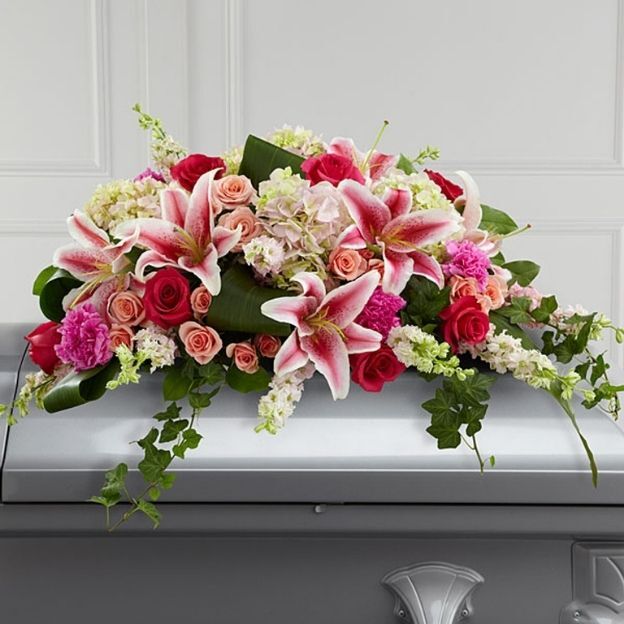 Casket spray with pink lilies, roses, and carnations in shades of pink, red, and peach, accented with greenery.