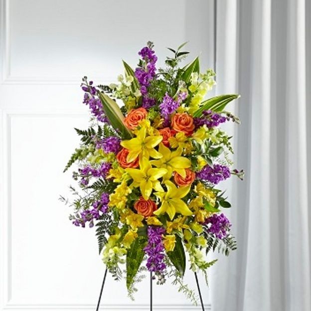 Standing funeral spray with yellow lilies, orange roses, purple stock, and greenery on a black easel.