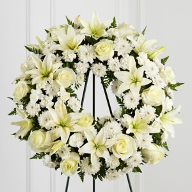 Circular floral wreath featuring white blooms and greenery, displayed on a tripod stand for a memorial service.