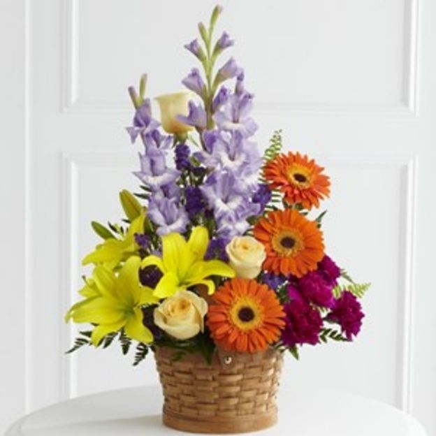 Mixed bouquet in a basket featuring yellow lilies, orange gerberas, purple delphiniums, and cream roses.