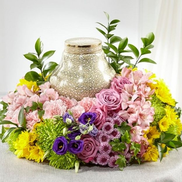 Elegant urn surrounded by a vibrant floral wreath of pink roses, yellow daisies, purple lisianthus, and green mums.