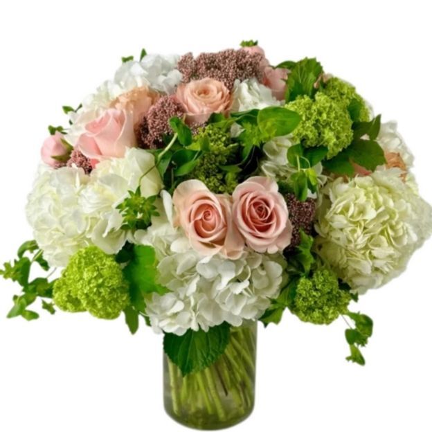 Mixed floral arrangement with soft pink roses, fluffy white blooms, and vibrant green accents in a clear vase.