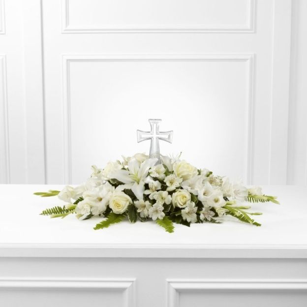 Elegant white floral display featuring lilies and roses with a silver cross, set against a white-paneled background.