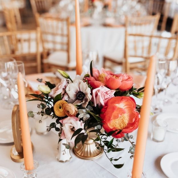 Elegant wedding table with peach candles, gold vase, and a vibrant floral centerpiece of peonies and ranunculus.