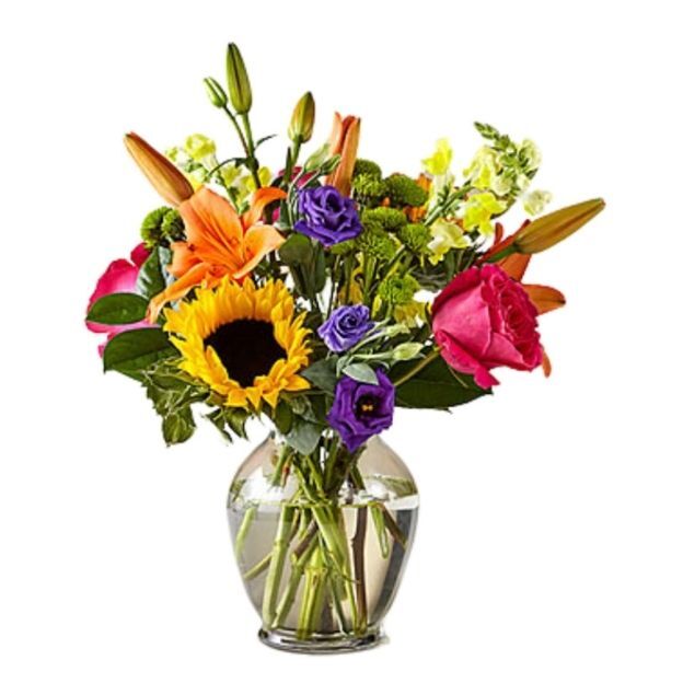 Colorful mixed flower arrangement featuring sunflowers, roses, and lilies in a round glass vase.