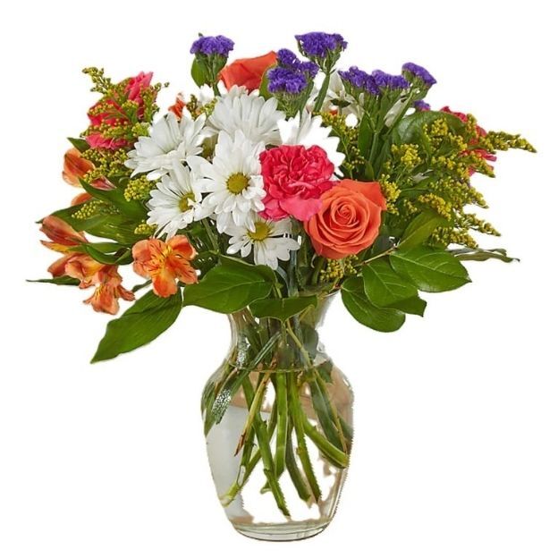 A vibrant flower bouquet in a clear glass vase featuring roses, daisies, carnations, and greenery.