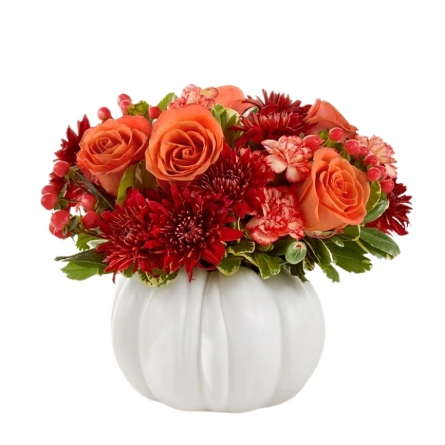 Autumn bouquet of orange and red flowers with pink carnations and greenery in a white pumpkin-shaped container.