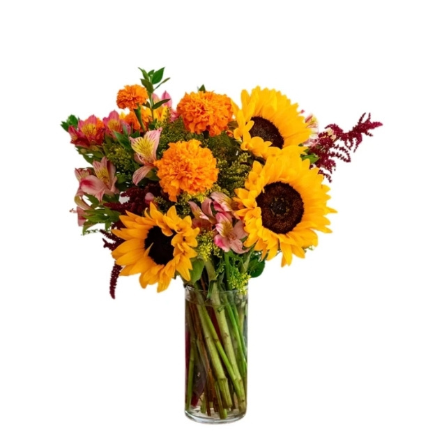 Yellow sunflowers, orange marigolds, pink flowers, and burgundy accents in a tall clear vase with visible stems.