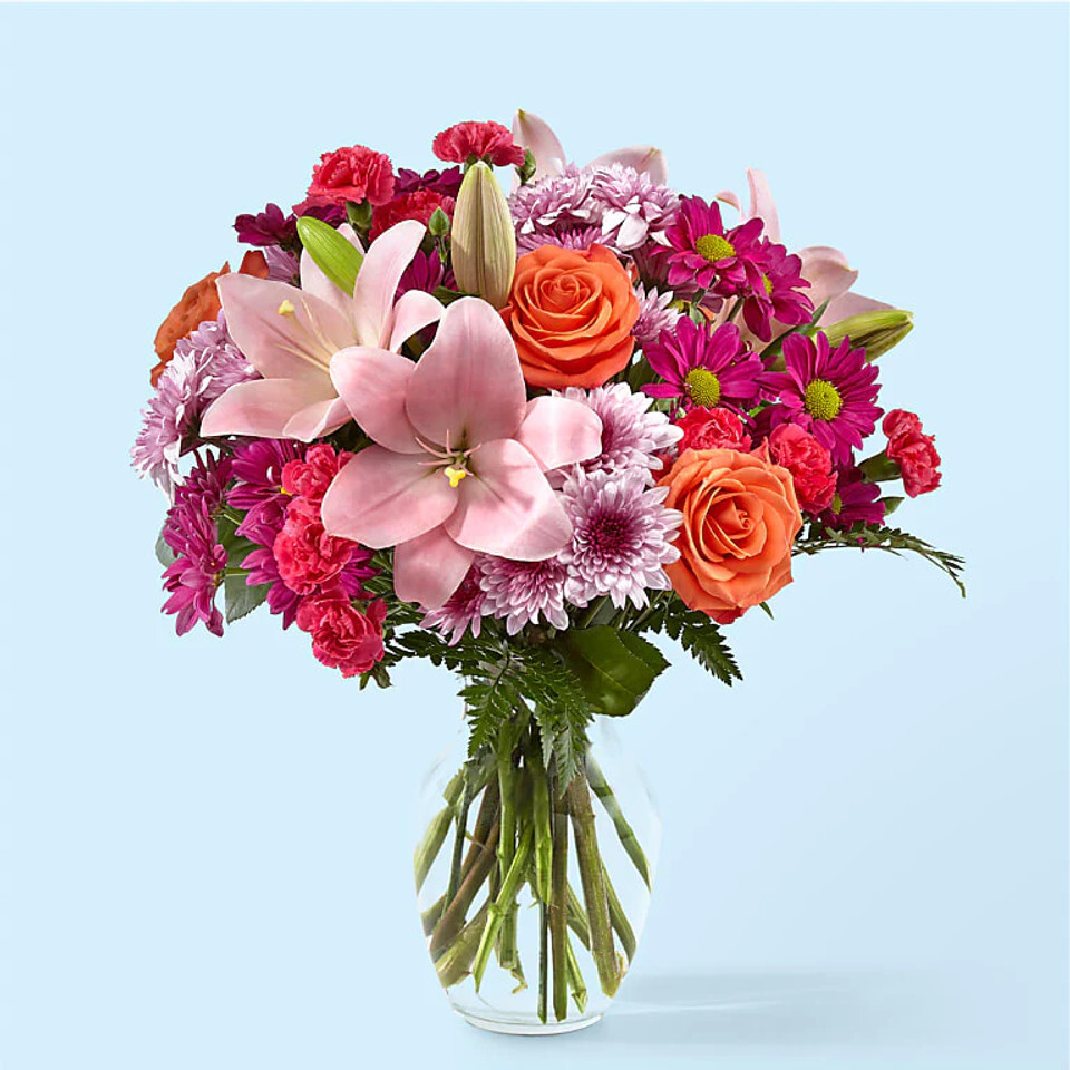 Vibrant bouquet with pink lilies, orange roses, purple daisies, and carnations in a clear glass vase.